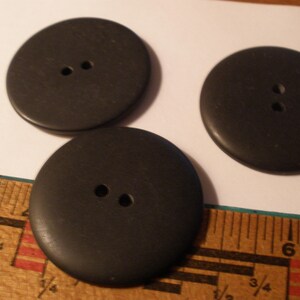 May include: Three large, round, black buttons with two holes each. The buttons are made of a solid material and are lying on a white surface next to a wooden ruler.