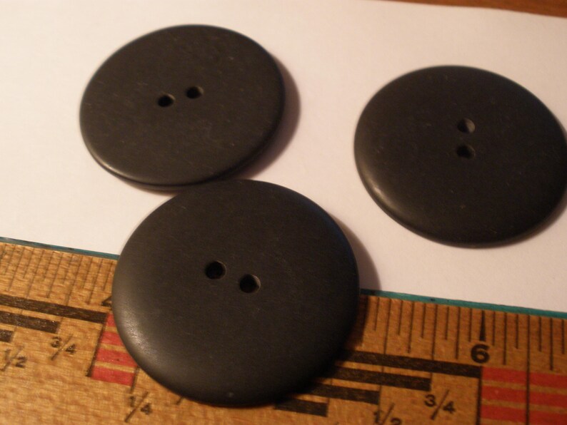 May include: Three large, round, black buttons with two holes each. The buttons are made of a solid material and are lying on a white surface next to a wooden ruler.