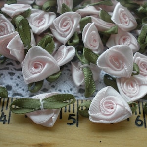 May include: A collection of small, pink satin roses with green ribbon leaves. The roses are arranged on a white lace background with a wooden ruler in the foreground.