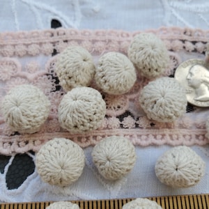 May include: A collection of 10 off-white crocheted buttons. The buttons are round and have a raised, textured design. They are arranged on a pink and white lace fabric.