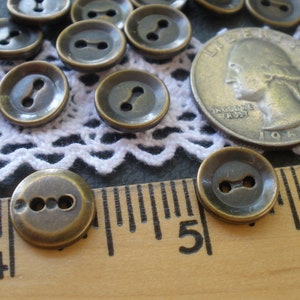 May include: A collection of 15 antique brass buttons with two holes each. The buttons are arranged on a white lace doily and a wooden ruler.