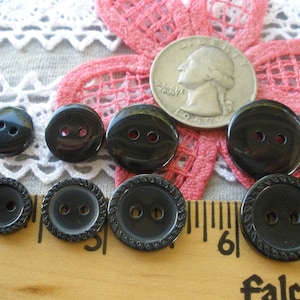 May include: Eight black buttons with two holes each. Some buttons have a decorative edge. The buttons are arranged on a wooden ruler and a piece of fabric. A US quarter is included for size comparison.