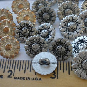May include: A collection of silver and gold metal daisy buttons with a raised design. The buttons are arranged on a wooden ruler with markings in inches.