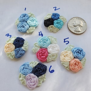 May include: Six handmade fabric flower embellishments in various colors, including blue, pink, white, and cream. Each flower is made of ribbon and has a layered, rosette-like appearance. The flowers are arranged in a circle on a white surface.