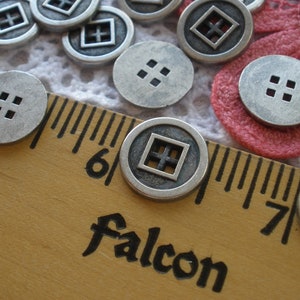 May include: Close-up of several silver-colored metal buttons. Some buttons have a square design with a cross, while others have four square holes. A wooden ruler with the word "Falcon" is in the foreground.