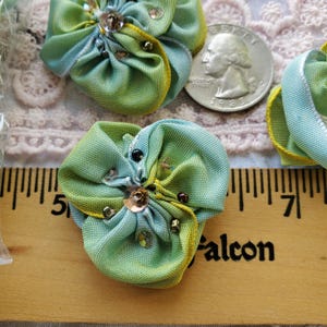 May include: Two green and blue fabric flowers with sequins and beads. The flowers are about 2 inches in diameter and are sitting on a wooden ruler.