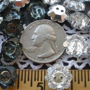 May include: A close-up of a collection of clear and dark gray flower-shaped buttons. The buttons are arranged on a white lace fabric with a quarter and a ruler for scale.  The buttons are approximately 1/2 inch in diameter.