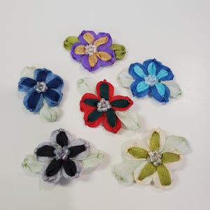 May include: A collection of six fabric flower embellishments in various colors. The flowers feature layered petals in shades of blue, purple, red, black, and cream, each with a central bead. Green and light green leaves are attached to the base of each flower.