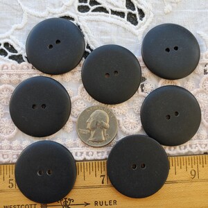 May include: Seven large, matte black buttons with two holes each, arranged on a lace surface. A U.S. quarter is included for size comparison. The buttons are round and appear to be made of a solid material.