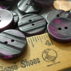 K&ouml;nnte beinhalten: Eine Sammlung von purpurfarbenen und schwarzen gestreiften Kn&ouml;pfen mit jeweils zwei L&ouml;chern. Die Kn&ouml;pfe sind auf einem Holzlineal mit dem aufgedruckten Text "Goose Shoes" angeordnet.