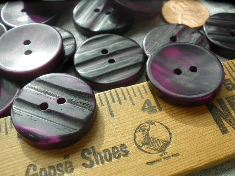 K&ouml;nnte beinhalten: Eine Sammlung von purpurfarbenen und schwarzen gestreiften Kn&ouml;pfen mit jeweils zwei L&ouml;chern. Die Kn&ouml;pfe sind auf einem Holzlineal mit dem aufgedruckten Text "Goose Shoes" angeordnet.