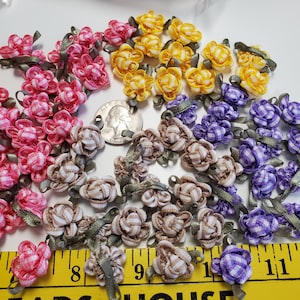 May include: A collection of small fabric ribbon roses in pink, yellow, purple, and beige gingham patterns, each with a tiny green leaf. A US quarter coin and a yellow measuring tape showing inches are placed among the flowers for scale reference.