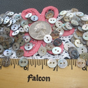 May include: A collection of approximately 100 vintage mother of pearl and wood buttons in various shades of gray, brown, and white. The buttons are arranged on a white lace doily with a pink flower in the center. A US quarter is included for scale.
