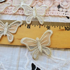 May include: Delicate, embroidered butterfly appliques in gold and cream. The butterflies are arranged on a white surface with a wooden ruler showing measurements in inches. The wings have a sheer, mesh-like appearance.