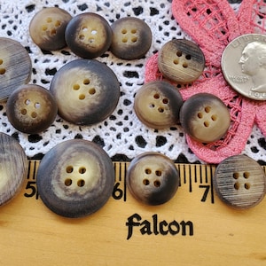 May include: A collection of brown and tan buttons with four holes, in various sizes. The buttons have a marbled appearance and are arranged on a white lace doily and a wooden ruler. The word "Falcon" is printed on the ruler.