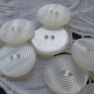 May include: A collection of white, pearlescent buttons with a spiral design. The buttons have two or four holes for sewing and are arranged on a textured, white fabric. The buttons vary in size and shape.