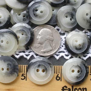 May include: A collection of white and black marbled buttons with two holes each. The buttons are arranged on a white lace doily and a wooden ruler.