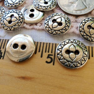 May include: Silver-toned metal buttons with a decorative floral design and two holes. The buttons are arranged on a light pink lace fabric and a wooden ruler. A US quarter is also visible.
