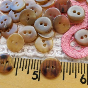 May include: A collection of round, two-hole brown buttons with a mottled pattern. A wooden ruler is positioned at the bottom, showing the buttons measure approximately 0.5 inches in diameter. The buttons are scattered across a textured surface.