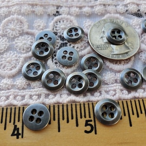 May include: A collection of small, round, gunmetal-colored buttons with four holes, arranged on a lace surface. A ruler with inch markings and a US quarter are also present, providing scale. The buttons have a slightly aged appearance.