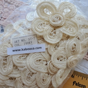May include: Ivory-colored lace trim with a floral design. The trim is made of a delicate, woven material and is packaged in a clear plastic bag. The label reads "ART NO. ST-7 COL. IVORY" and "www.kaboooco.com".
