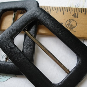 May include: Two black leather belt buckles with a silver metal buckle. The buckles are on a wooden ruler with the number 4 visible.
