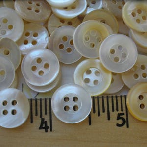 May include: A collection of white and cream colored pearl buttons with four holes each. The buttons are arranged on a wooden surface with a ruler.