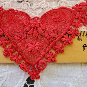 May include: A vibrant red lace heart with intricate floral and swirl details. The heart shape is the focal point, surrounded by delicate flower-shaped embellishments. The lace is set against a neutral background, highlighting the detailed craftsmanship.