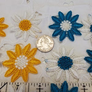 May include: A collection of embroidered flower patches in white, yellow, and blue, arranged around a United States quarter coin. The patches feature detailed stitching and are positioned above a ruler showing measurements in inches.