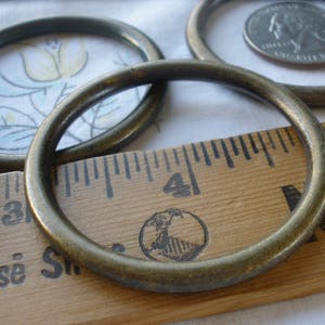 May include: Four antique brass rings of varying sizes, with one ring lying on top of a wooden ruler marked with inches. The ruler has the words "Goose" and "New" printed on it.