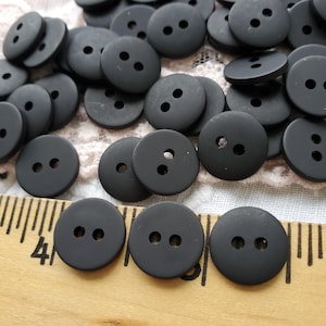 May include: A large group of black plastic buttons with two holes each. The buttons are arranged in a random pattern on a white surface.