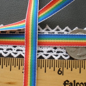 May include: Two rainbow striped ribbons, each with red, orange, yellow, green, blue, and indigo stripes, are laid out on a white crocheted surface. A ruler is visible in the bottom of the image, with the numbers 4, 6, and 7 visible.