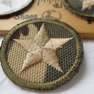 May include: A round, embroidered patch with a beige five-pointed star on a green and brown camouflage background.
