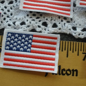 May include: A set of four small American flag patches. The patches are made of fabric and have a white background with red and blue stripes. The stars are blue and white. The patches are about 2 inches by 3 inches.