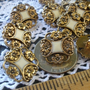 May include: Close-up of several vintage gold-tone buttons with a floral design and cream-colored center inlays. The buttons are arranged on a white lace surface, showcasing their intricate details and aged appearance. A few buttons are in focus, highlighting their ornate design.