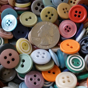 May include: A collection of colorful buttons in various sizes and shapes, including round and flat designs. The buttons are in shades of blue, green, yellow, orange, red, and pink. A US quarter coin is in the center of the buttons.