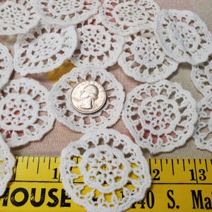 May include: A collection of white, handmade crochet doilies in various floral and circular designs. A U.S. quarter is placed on one of the doilies for size comparison. The doilies are arranged on a patterned surface, with a yellow ruler in the foreground.