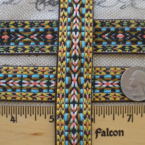 May include: Close-up of a colorful woven ribbon with a geometric pattern in shades of black, brown, gold, blue, green, and pink. The ribbon is approximately 1 inch wide and is measured against a ruler.