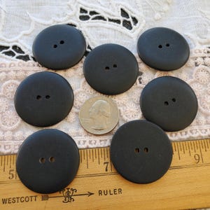 May include: A collection of seven large, matte black buttons with two holes each. A U.S. quarter is included for size comparison. The buttons are arranged on a wooden ruler, with the word "RULER" visible.