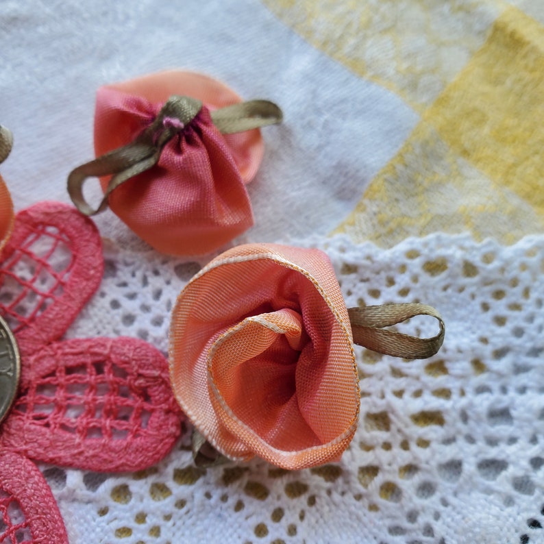 Op de afbeelding: Drie perzikkleurige bloemen van stof met bruine lintjes. De bloemen zijn gerangschikt op een wit kantonderleg.