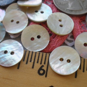 May include: A collection of 10 iridescent white mother of pearl buttons with two holes each. The buttons are arranged on a pink and white lace fabric with a wooden ruler in the foreground.