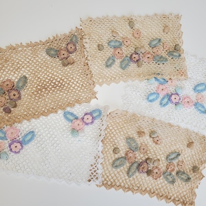May include: A collection of handmade crochet doilies in various shapes and sizes. The doilies feature delicate floral embroidery in pastel colors, including pink, blue, and beige. The crochet work has a vintage aesthetic.
