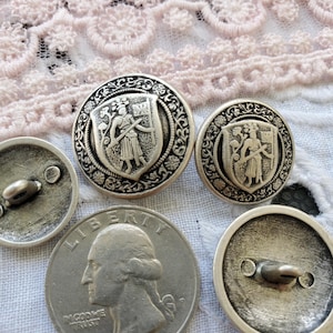 May include: Four silver-toned buttons with a raised design of a person in armor holding a sword and shield. The buttons have a decorative border and are approximately 1 inch in diameter.