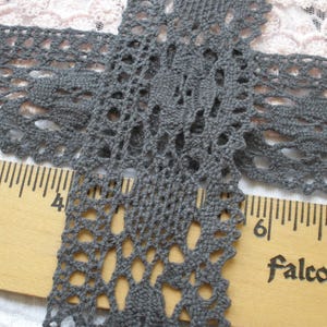 May include: Close-up of a length of dark gray lace with an open, floral design. The lace is laid over a wooden ruler with inch markings and the word "Falcon" printed on it. The lace is approximately 1.5 inches wide.