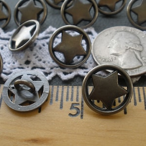 May include: A close-up of 10 silver-toned metal buttons with a five-pointed star design. The buttons are approximately 1 inch in diameter and have a shiny finish. The buttons are arranged on a white lace fabric background with a ruler for scale.