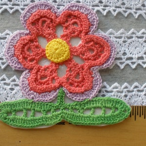 May include: A crocheted flower applique with a pink, orange, and yellow center, green leaves, and a white background.