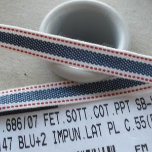 May include: Close-up of a white ribbon with a navy blue and red striped design. The ribbon has a woven texture and is likely made of cotton. The text on the label reads "ART.686/07 FET.SOTT.COT.PPT SB-V48 + C1147 BLU+2 IMPUN.LAT PL C.55 (F10)".