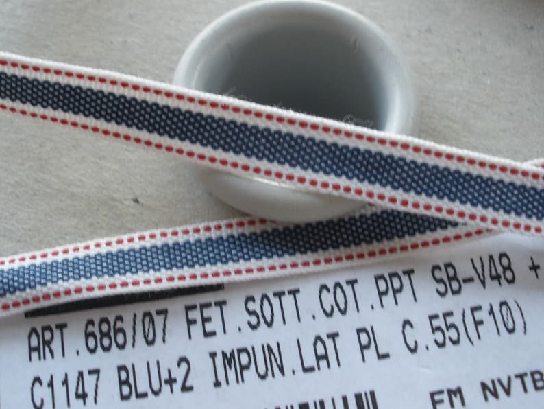 May include: Close-up of a white ribbon with a navy blue and red striped design. The ribbon has a woven texture and is likely made of cotton. The text on the label reads "ART.686/07 FET.SOTT.COT.PPT SB-V48 + C1147 BLU+2 IMPUN.LAT PL C.55 (F10)".