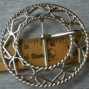 May include: Silver-toned metal belt buckle with a decorative design of hearts and a rope-like border. The buckle is approximately 5 cm in diameter.