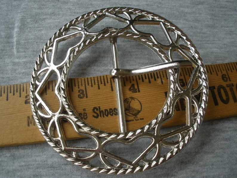 May include: Silver-toned metal belt buckle with a decorative design of hearts and a rope-like border. The buckle is approximately 5 cm in diameter.